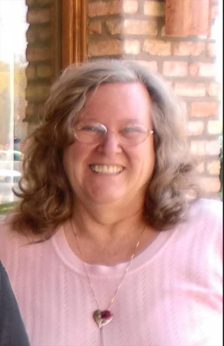Smiling person in a pink top and glasses, standing outdoors in front of a brick wall.