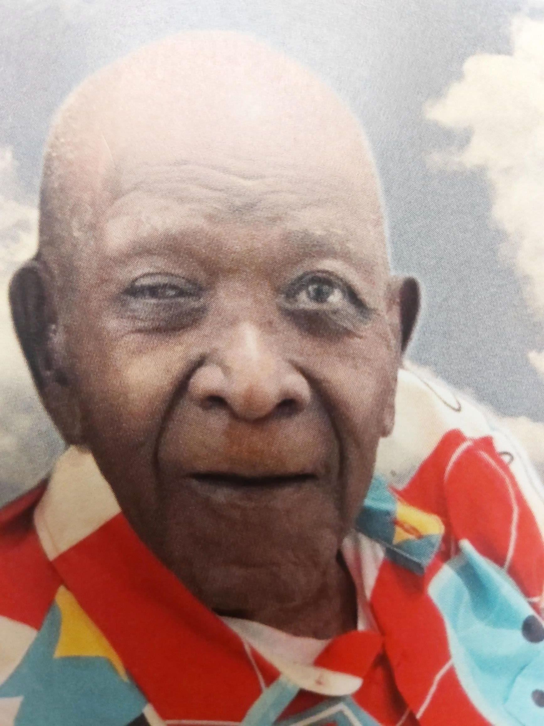 Close-up of an elderly man in a red and blue striped shirt, looking at the camera outdoors.