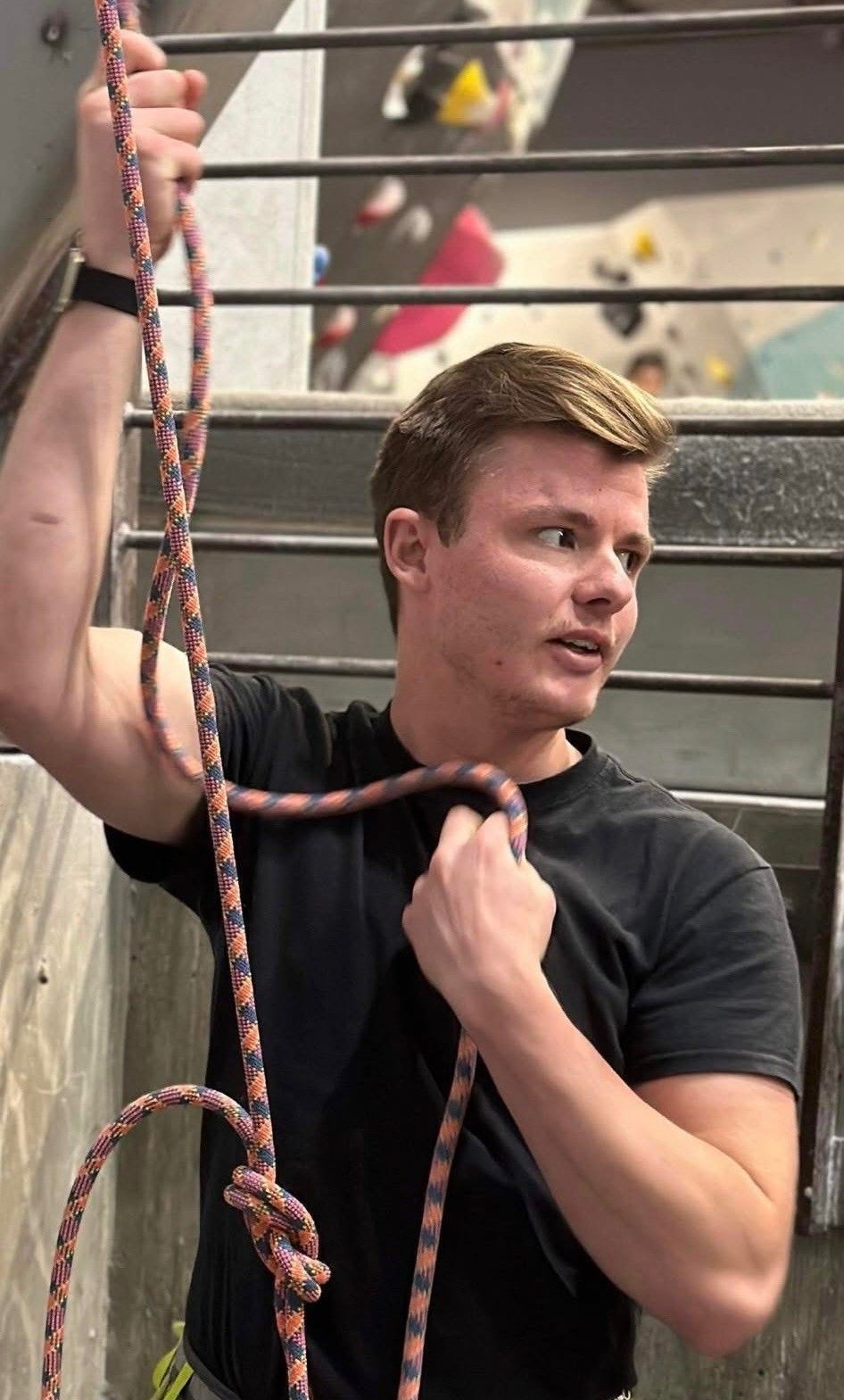Man in black shirt climbing rope indoors, holding the rope and looking to the side.