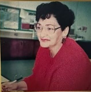 Woman in a red sweater and glasses seated at a desk, writing in a classroom or office setting