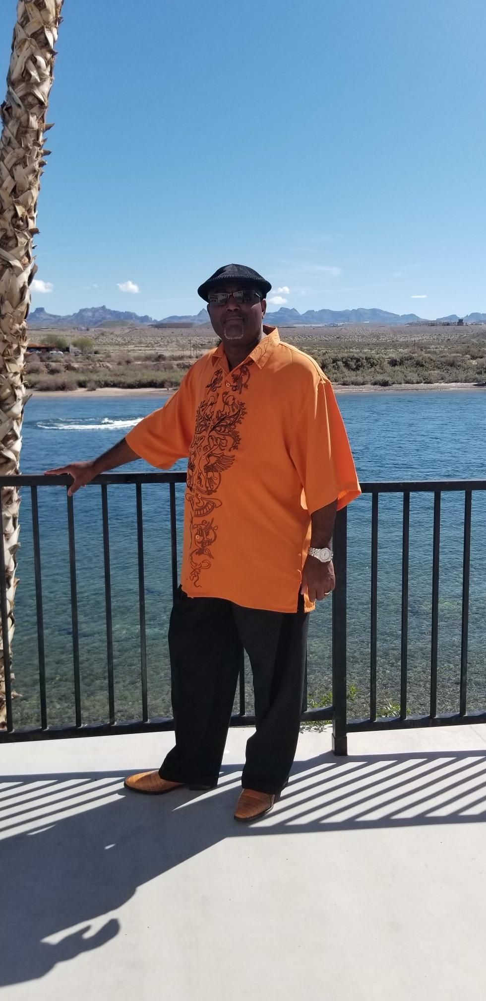Person in an orange shirt standing by a lakeside railing with mountains in the distance.