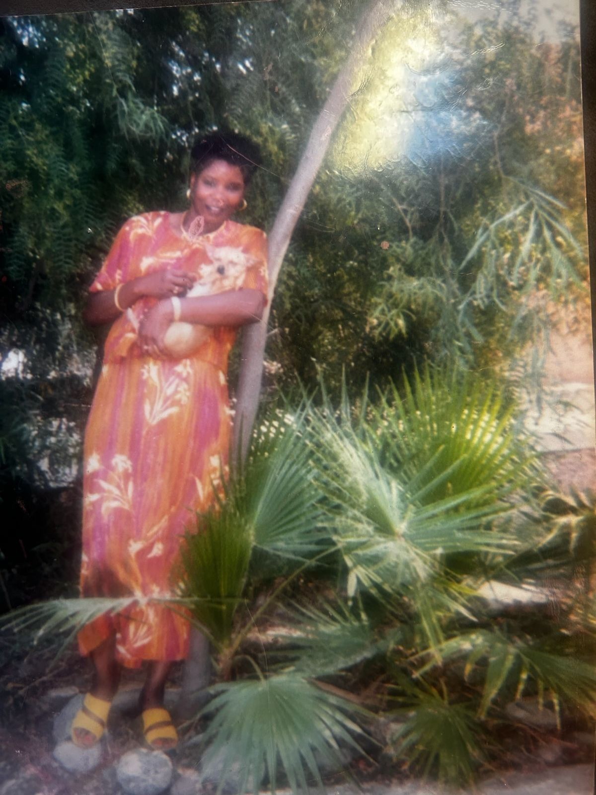 Person in an orange floral dress holding a baby beside a tree in a lush garden.