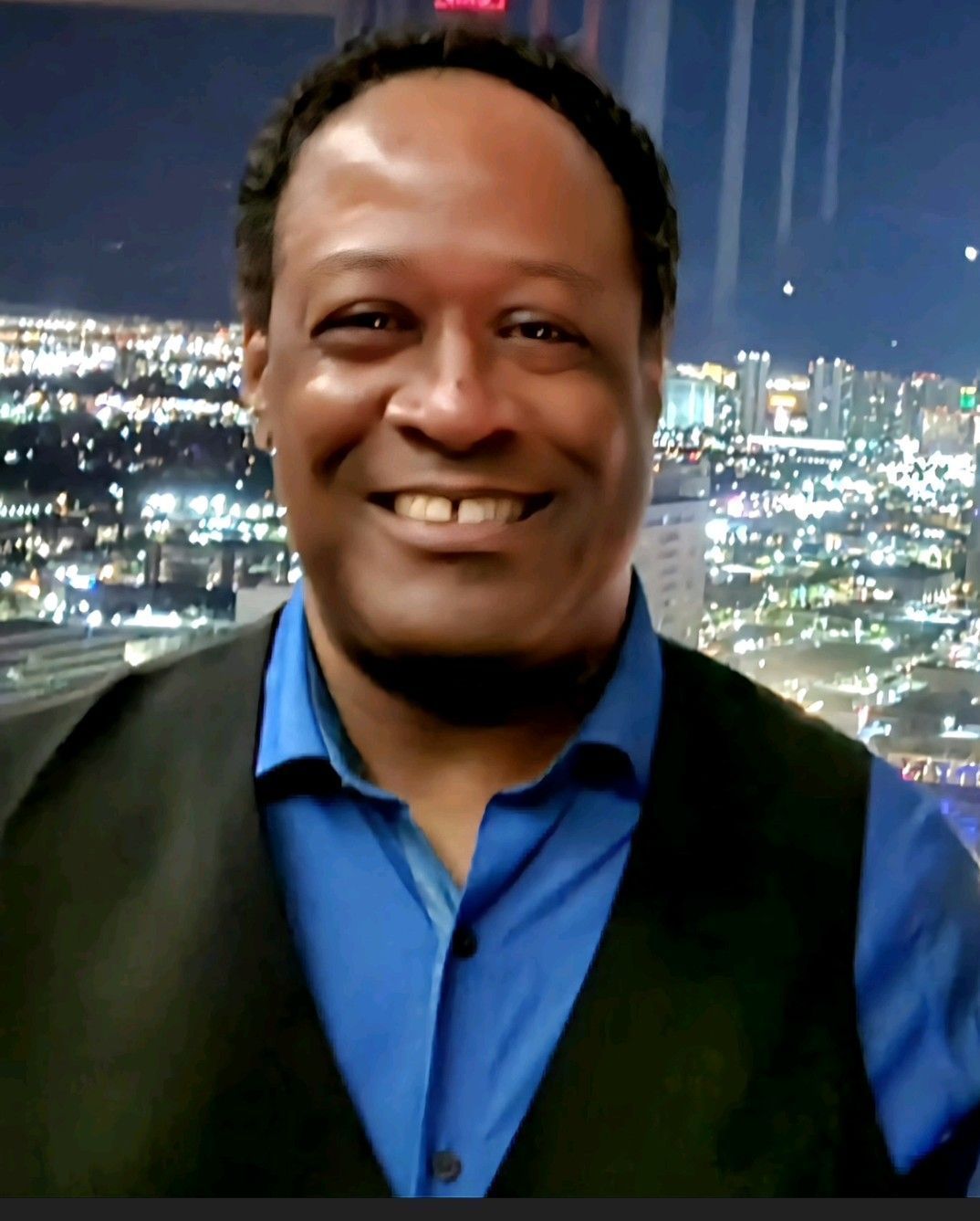 Smiling person in a blue shirt and black vest with a cityscape at night in the background