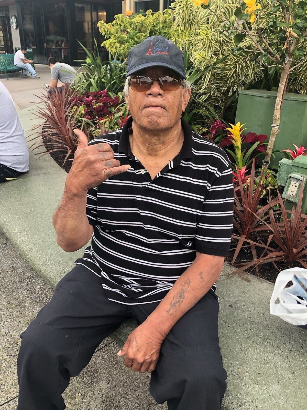 Man in a black striped shirt and cap sits outdoors, making a hand gesture in a garden setting
