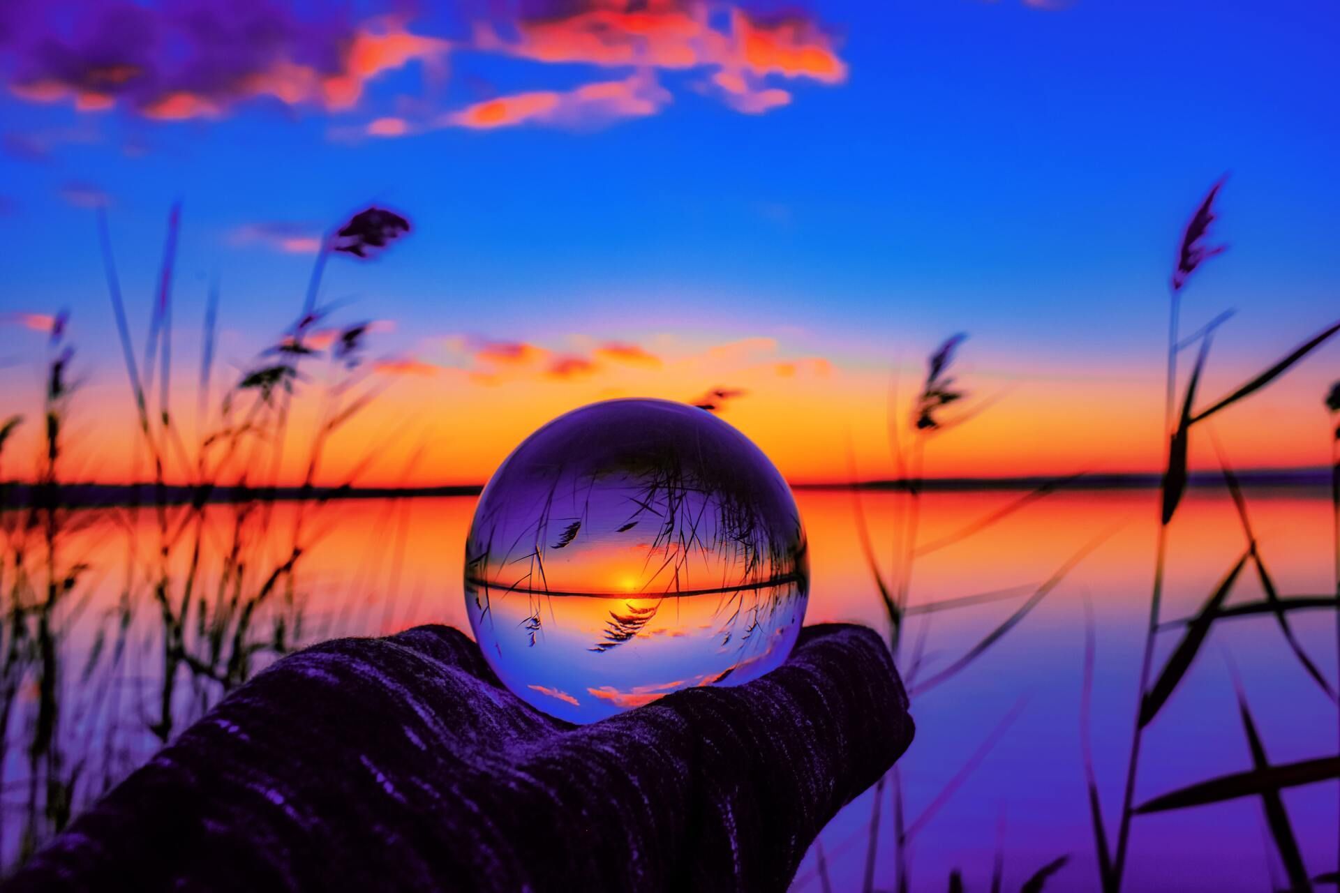 Glass orb reflecting a sunset over calm water, framed by silhouetted reeds and a purple-orange sky
