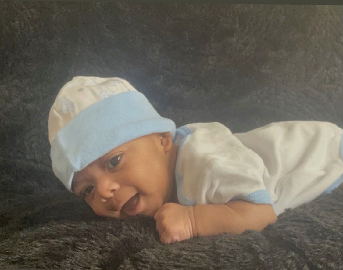 Baby in white and blue outfit lying on a dark blanket, looking toward the camera with tongue out