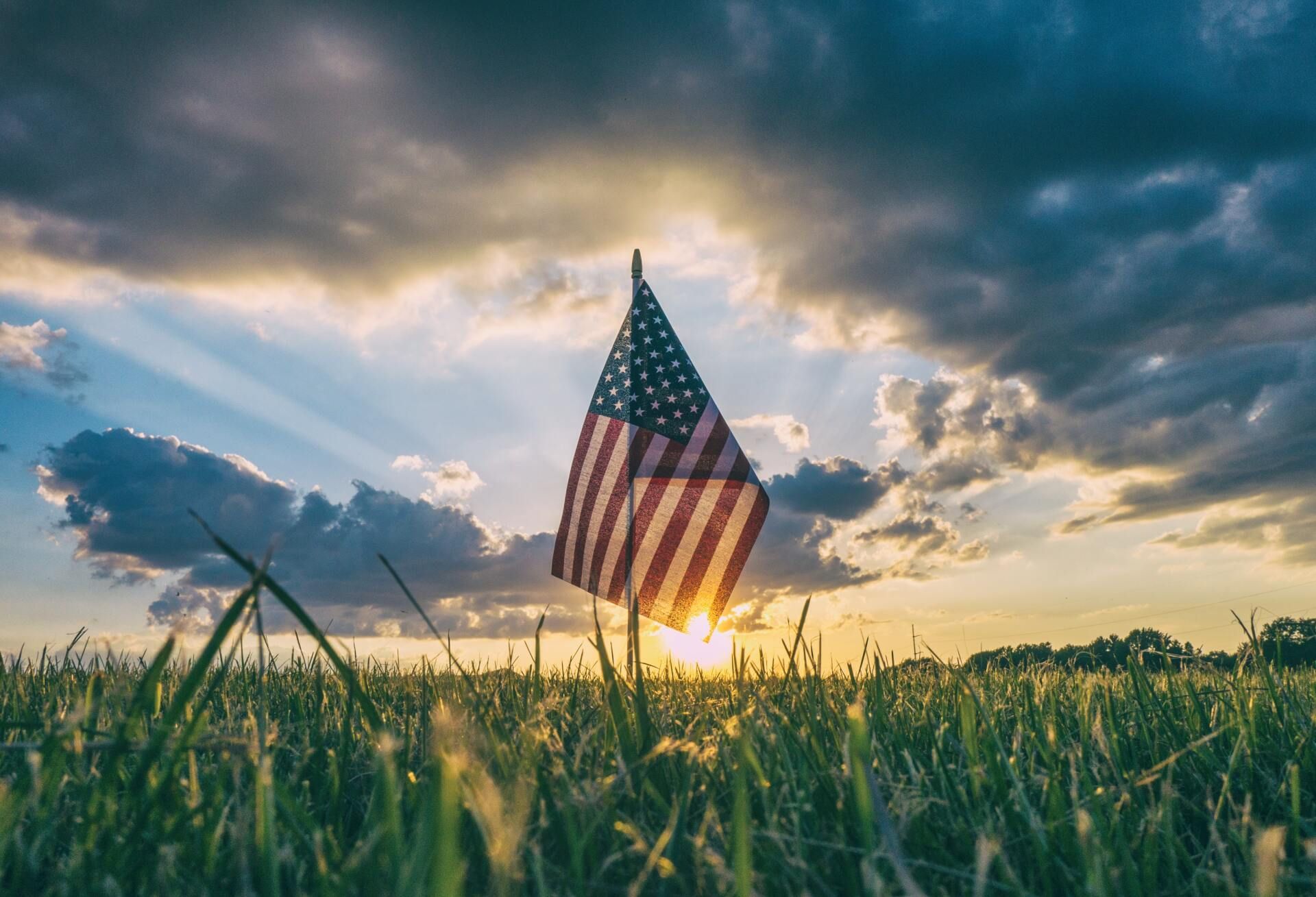 American flag planted in a grassy field at sunset under dramatic clouds