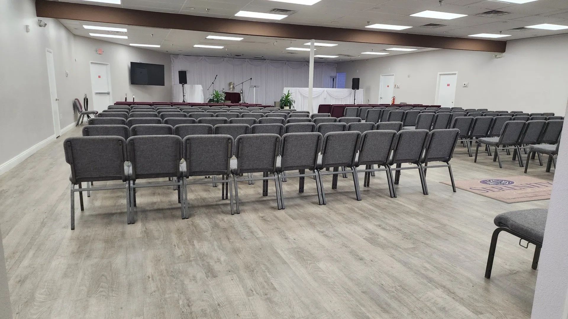 Empty conference room with rows of gray chairs facing the front, wood floor, and white walls.