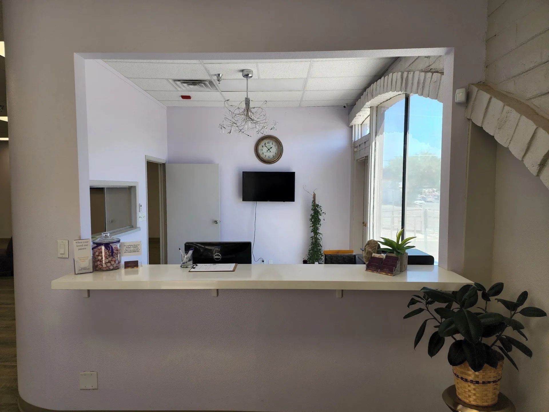 Living room view through a wide opening, with TV, wall clock, plants, and a bright window.