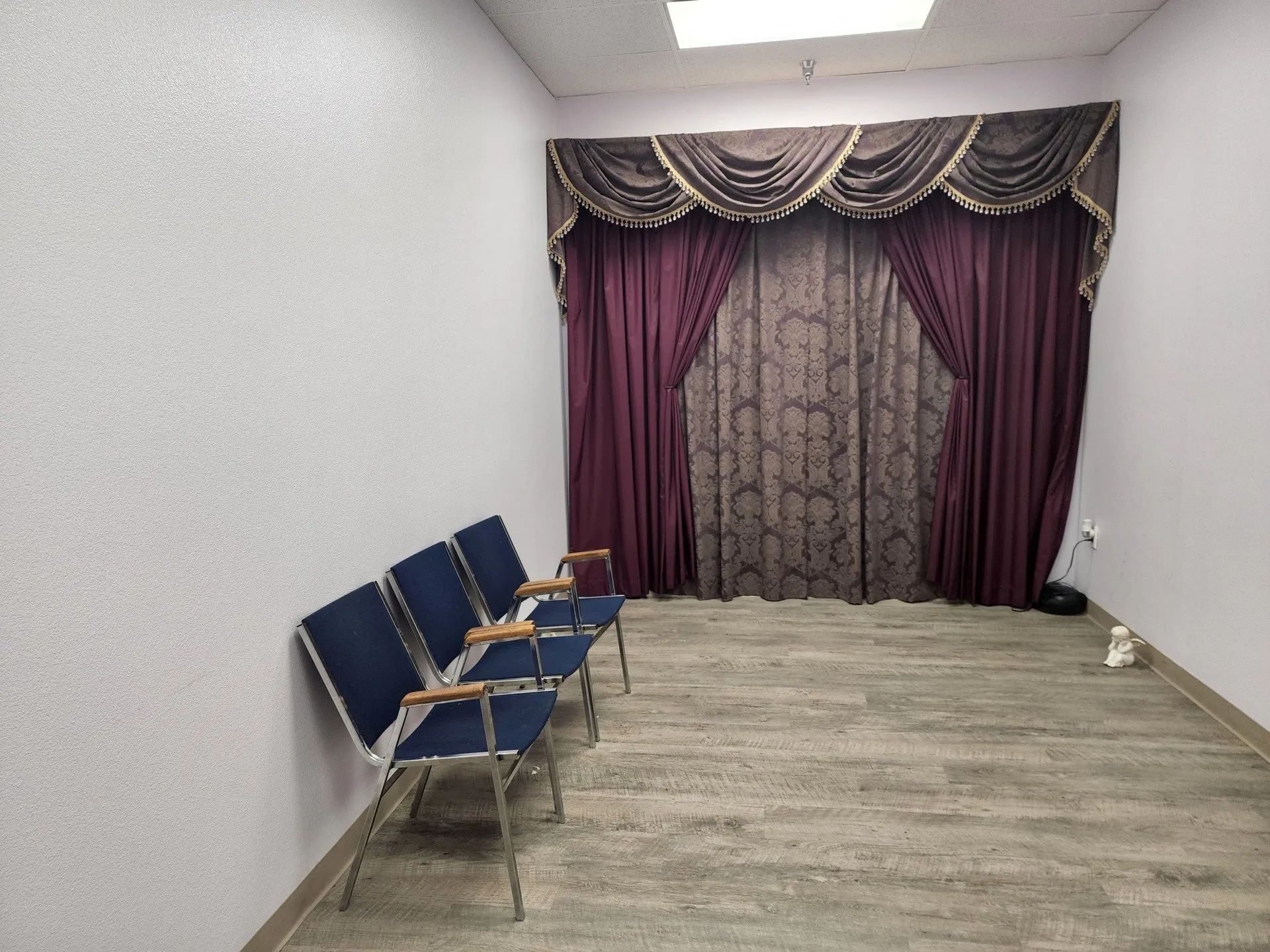 Small waiting room with blue chairs, wood floor, and purple curtains covering a window.