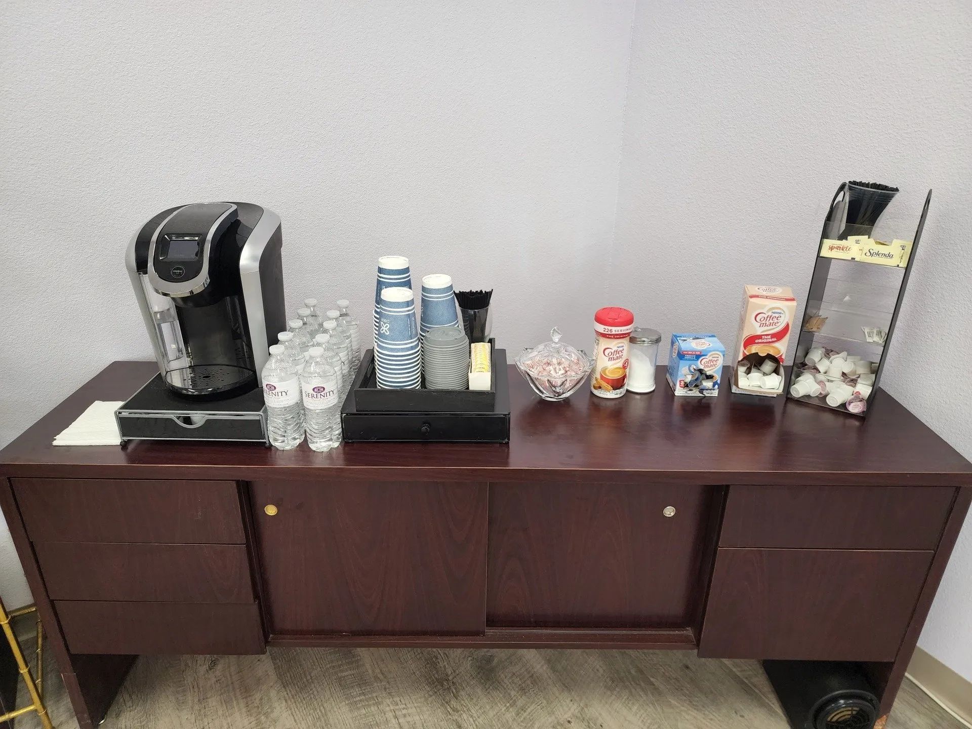 Coffee station with two machines and assorted cups, bottles, and snacks on a dark wooden cabinet