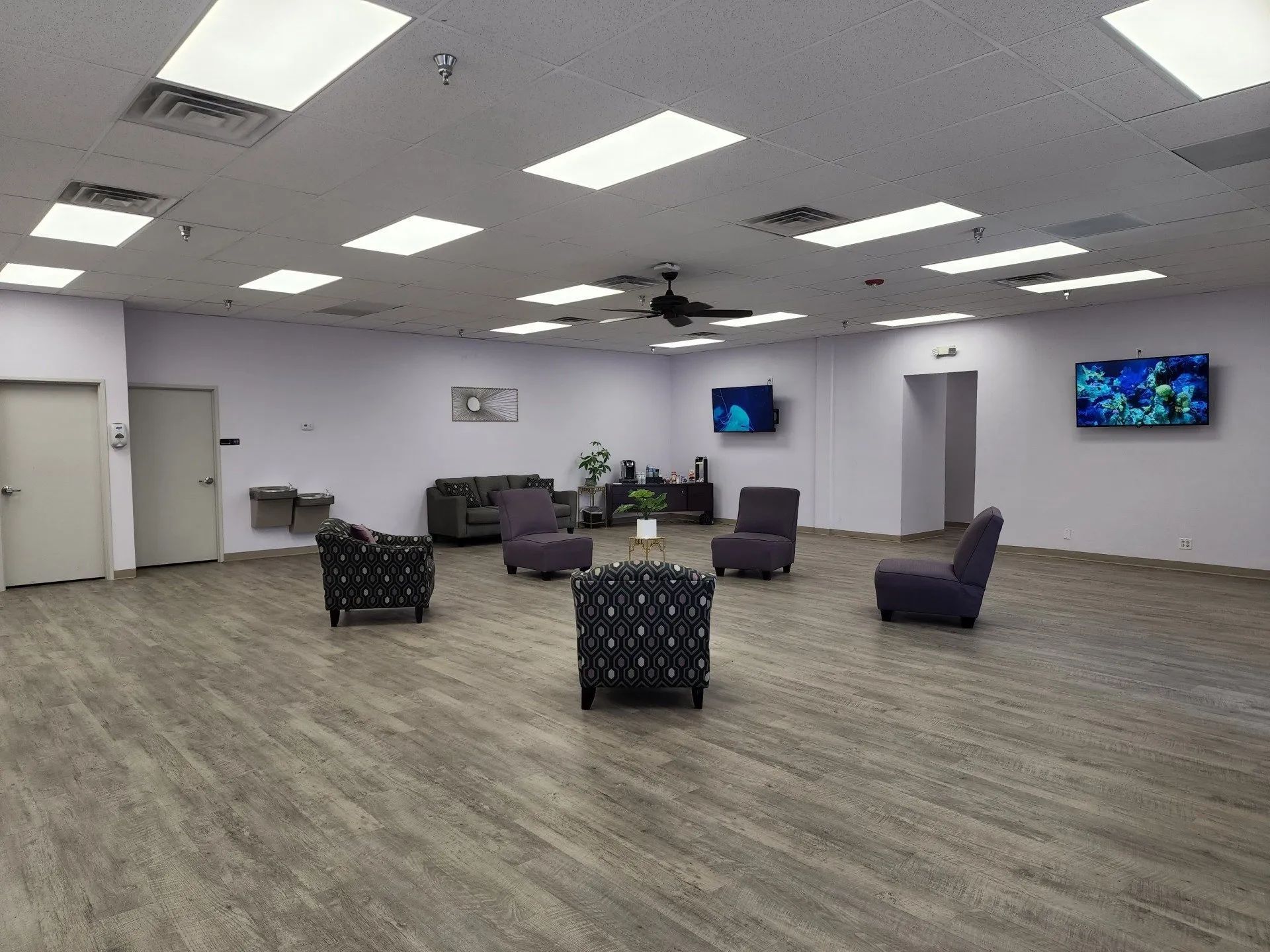 Spacious lounge with purple chairs, patterned armchairs, wall-mounted TVs, and bright overhead lights
