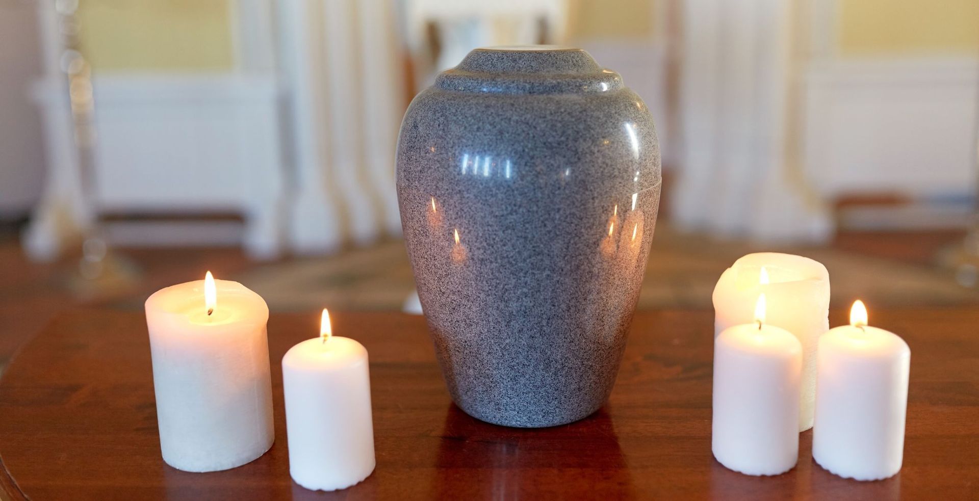 Large gray urn centered on a wooden table, flanked by lit white candles on both sides.