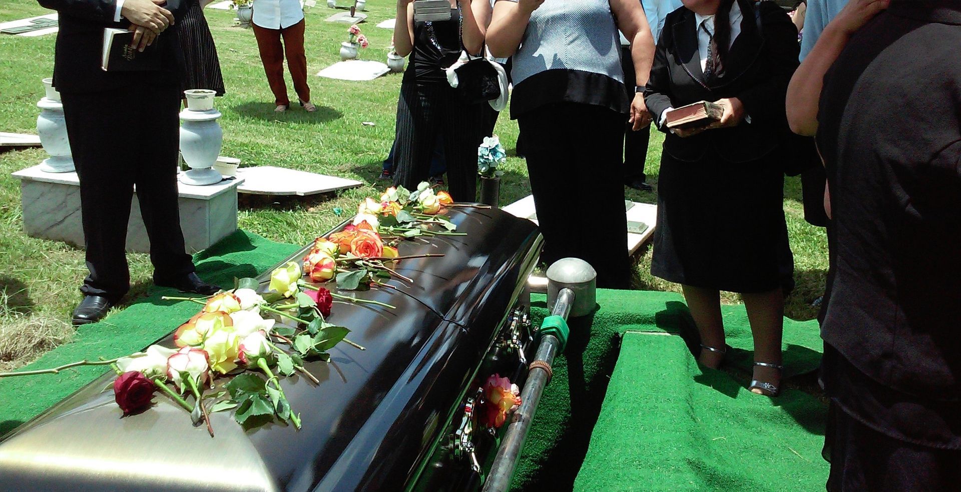 Mourners stand beside a flower-covered black casket on a green lawn during an outdoor funeral.