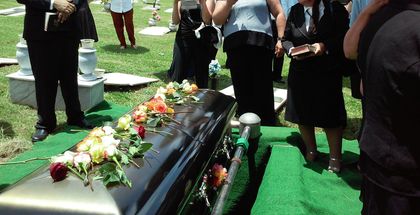 Mourners stand beside a flower-covered black casket on a green lawn during an outdoor funeral.