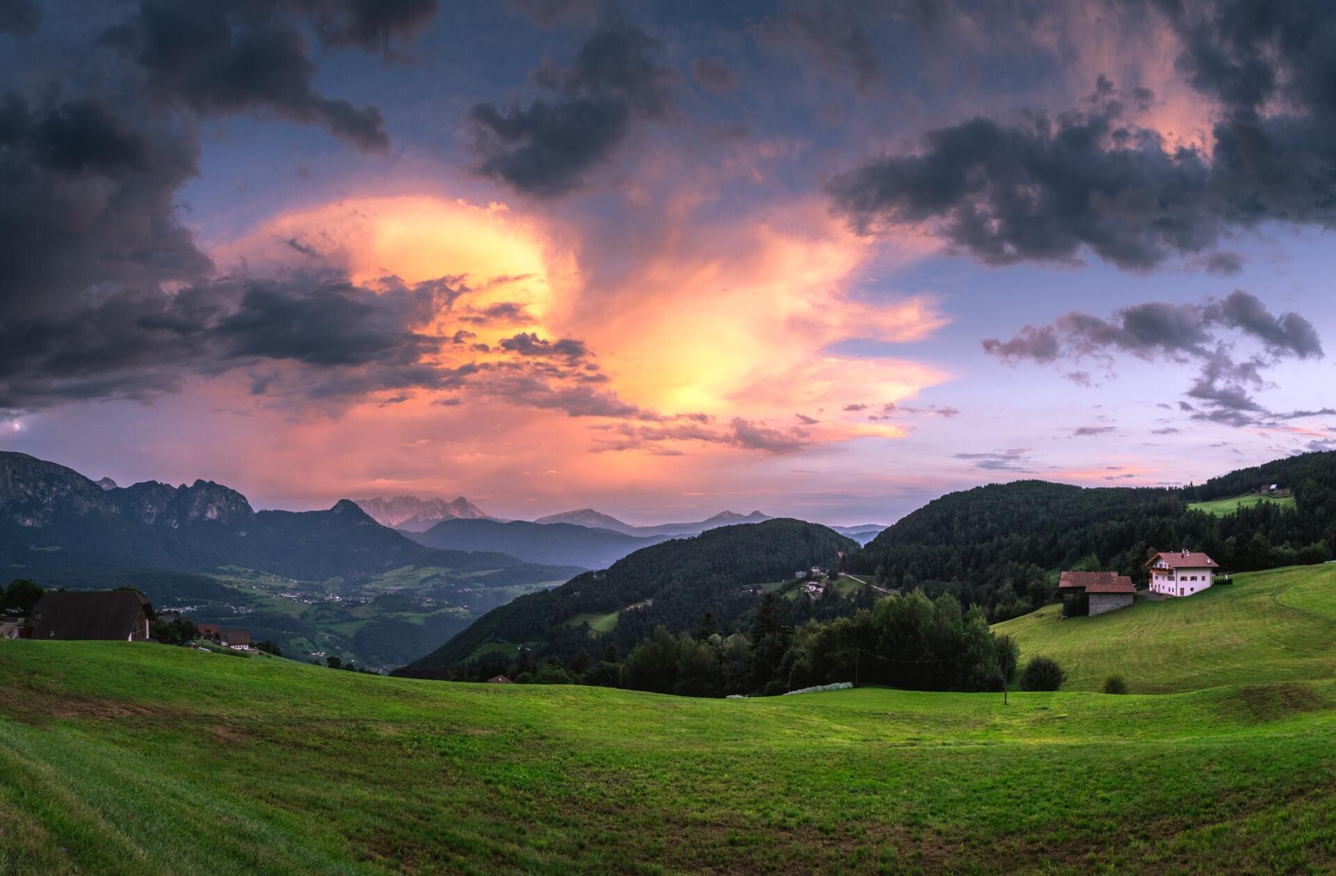 Sunset over green hills and fields, with a small house beneath dramatic purple clouds