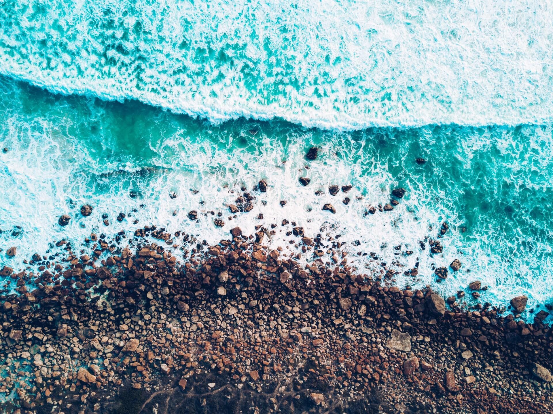 Turquoise waves breaking on a rocky shoreline, viewed from above.