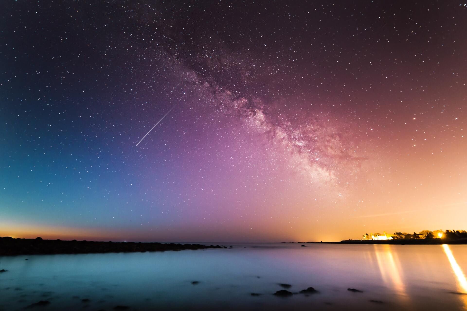 Starry night sky over calm water with a glowing purple-orange horizon