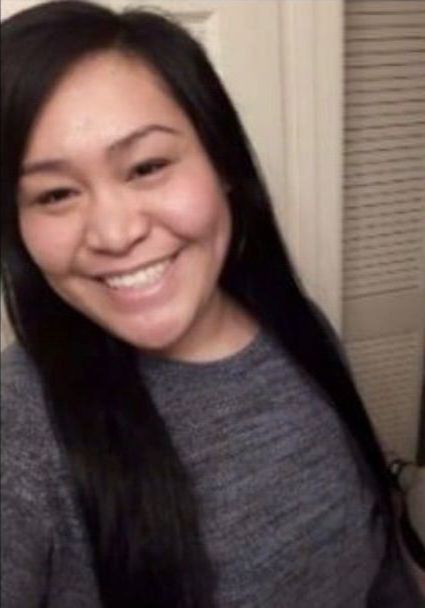 Smiling woman with long dark hair wearing a gray top, standing indoors near a door