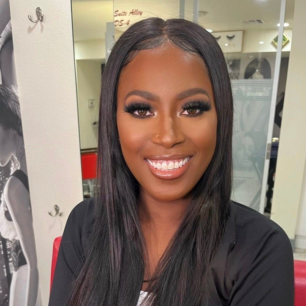 Smiling woman with long straight black hair in a black top, standing indoors at a salon.