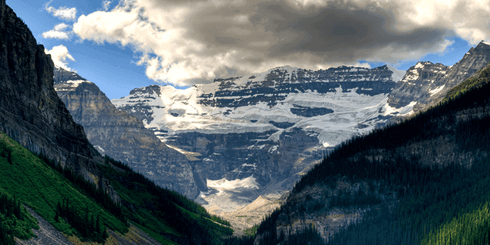 Snowy mountain range with a glacier under cloudy skies, framed by steep green valley cliffs