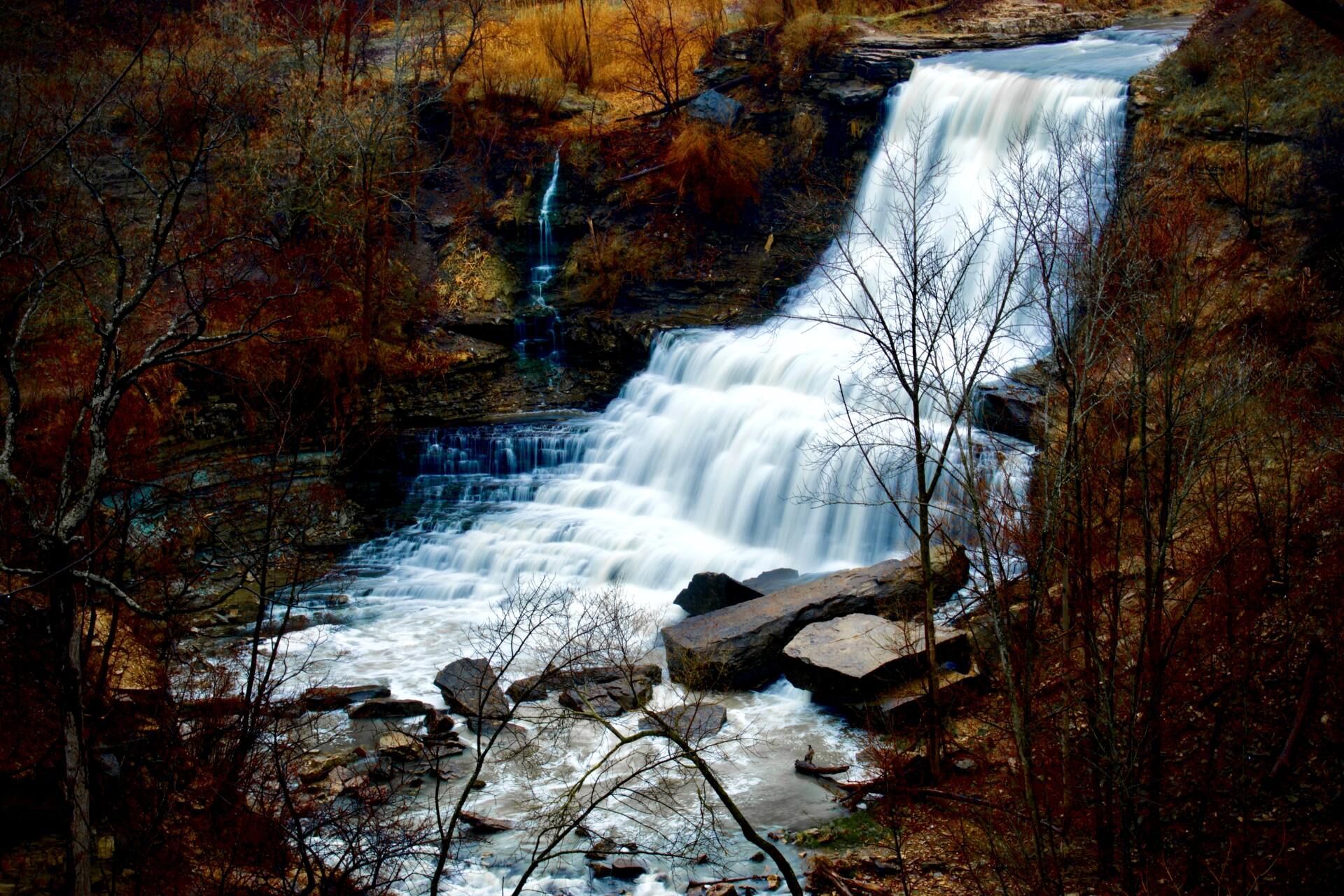 Waterfall cascading over dark rocks in a wooded ravine, with bare branches in the foreground.