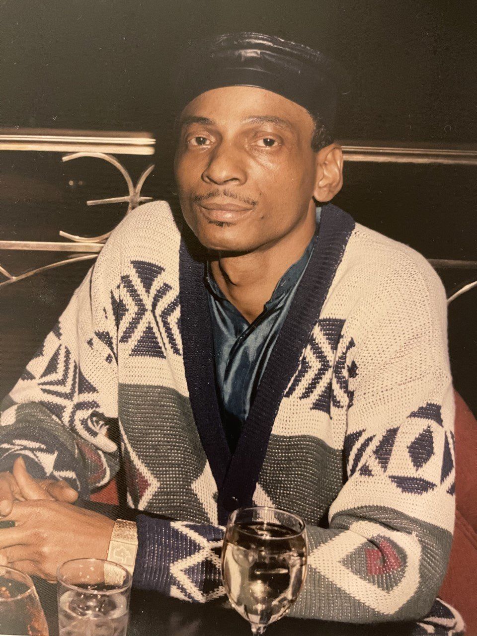 Man in a patterned cardigan seated at a table with a drink in a dimly lit restaurant.