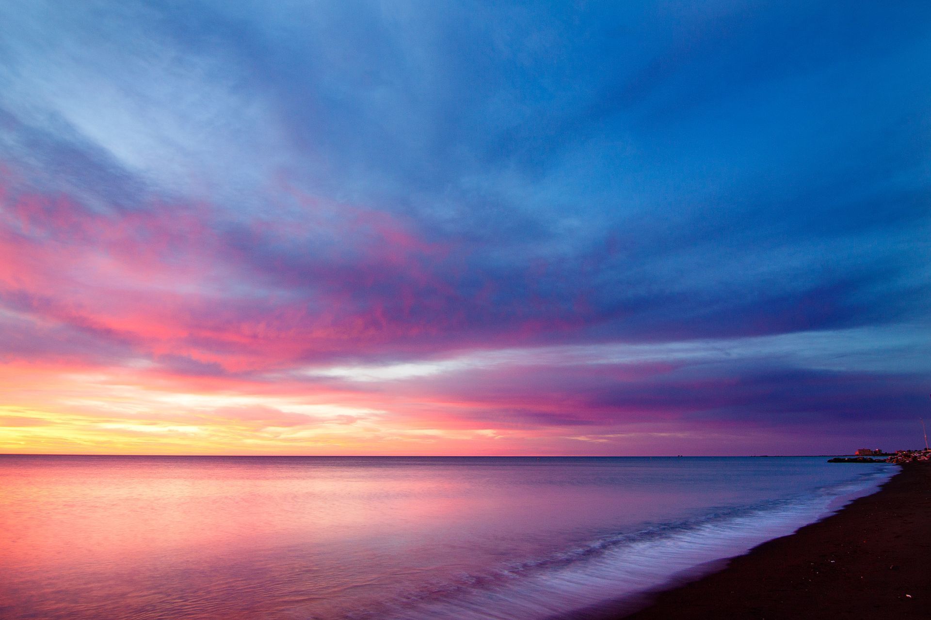 Colorful sunset over calm ocean with pink, purple, and blue clouds above a dark shoreline