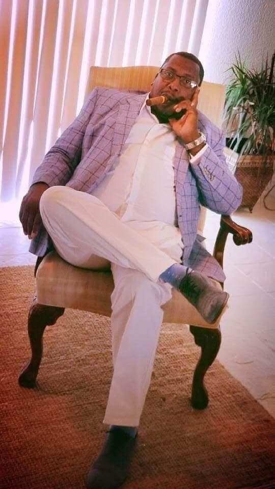 Man in a purple suit sitting cross-legged in a chair, holding a phone, with curtains and a plant behind him.