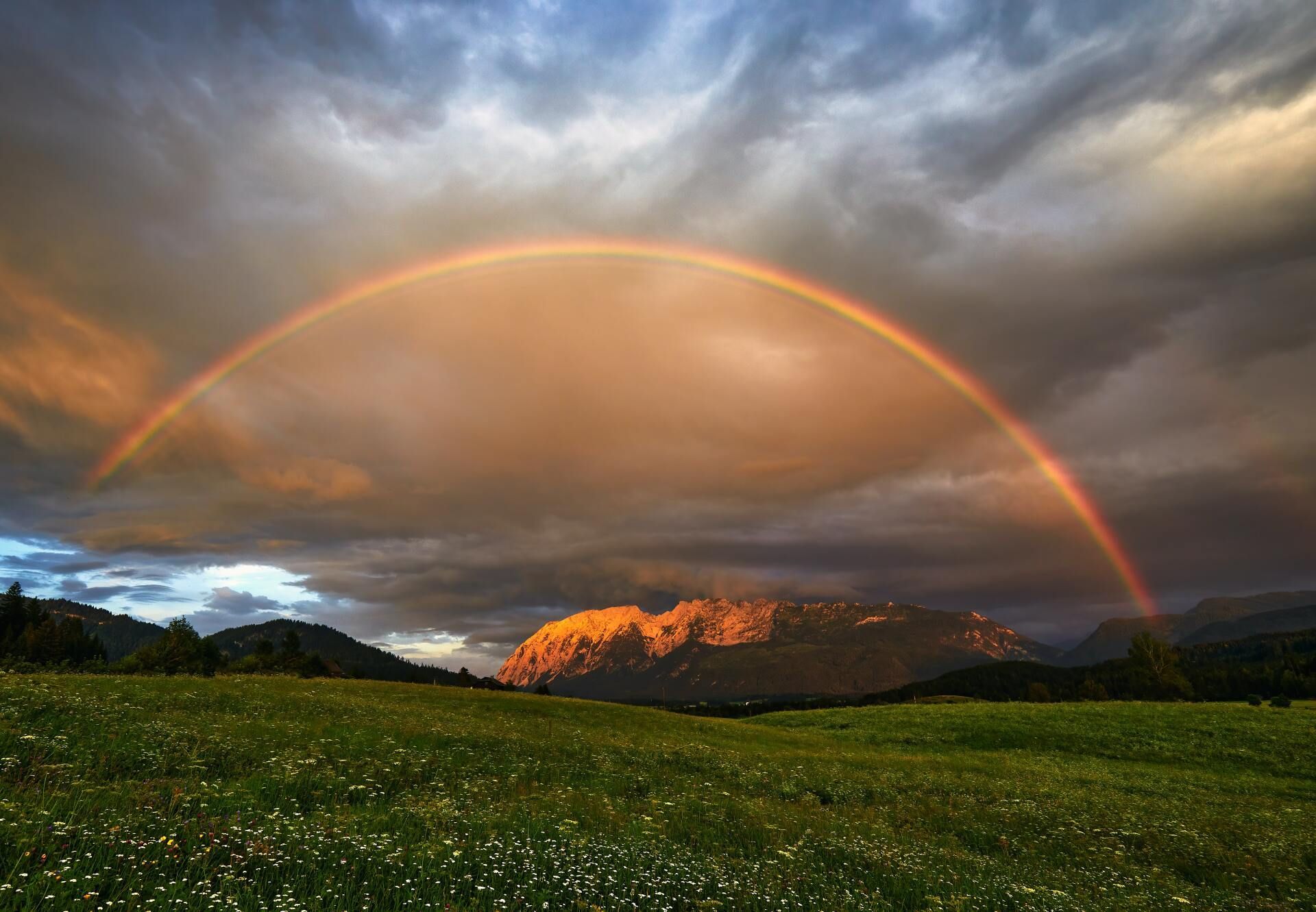 Rainbow over sunlit mountains and a green field under dramatic cloudy sky