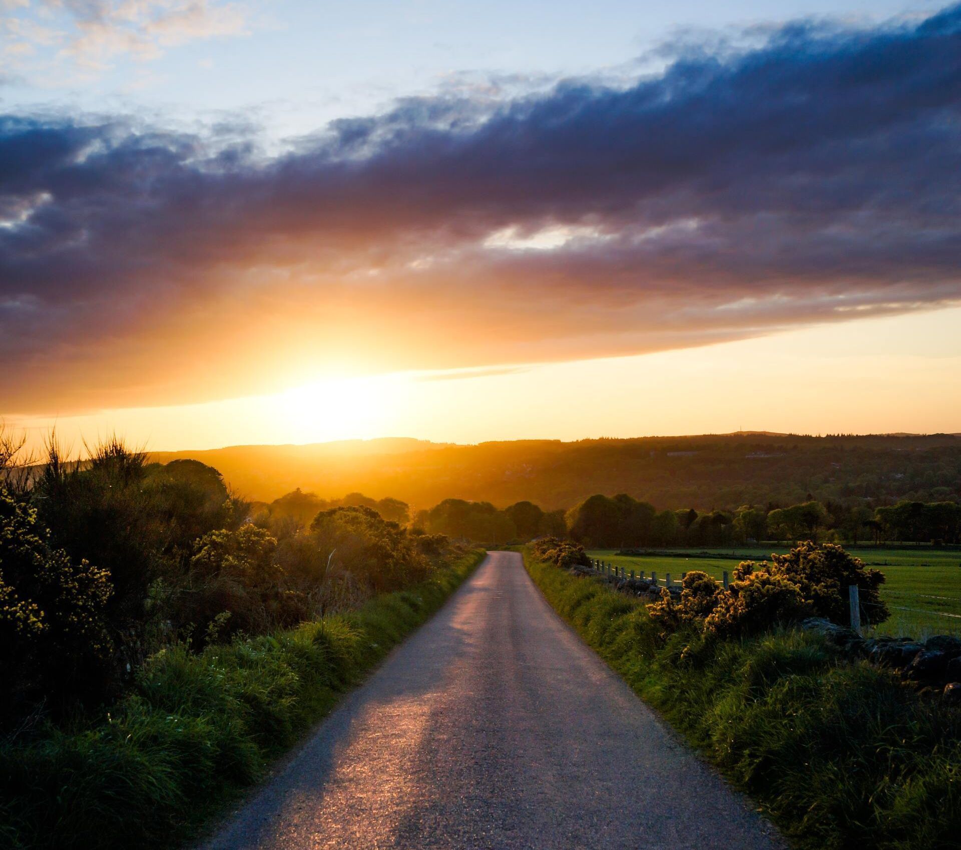 Sunset over a country road flanked by green fields and trees under a dramatic cloudy sky