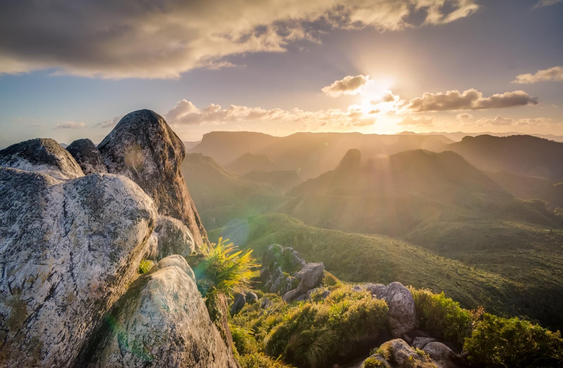 Sunlit mountain landscape with rocky foreground, green valleys, and clouds at sunset