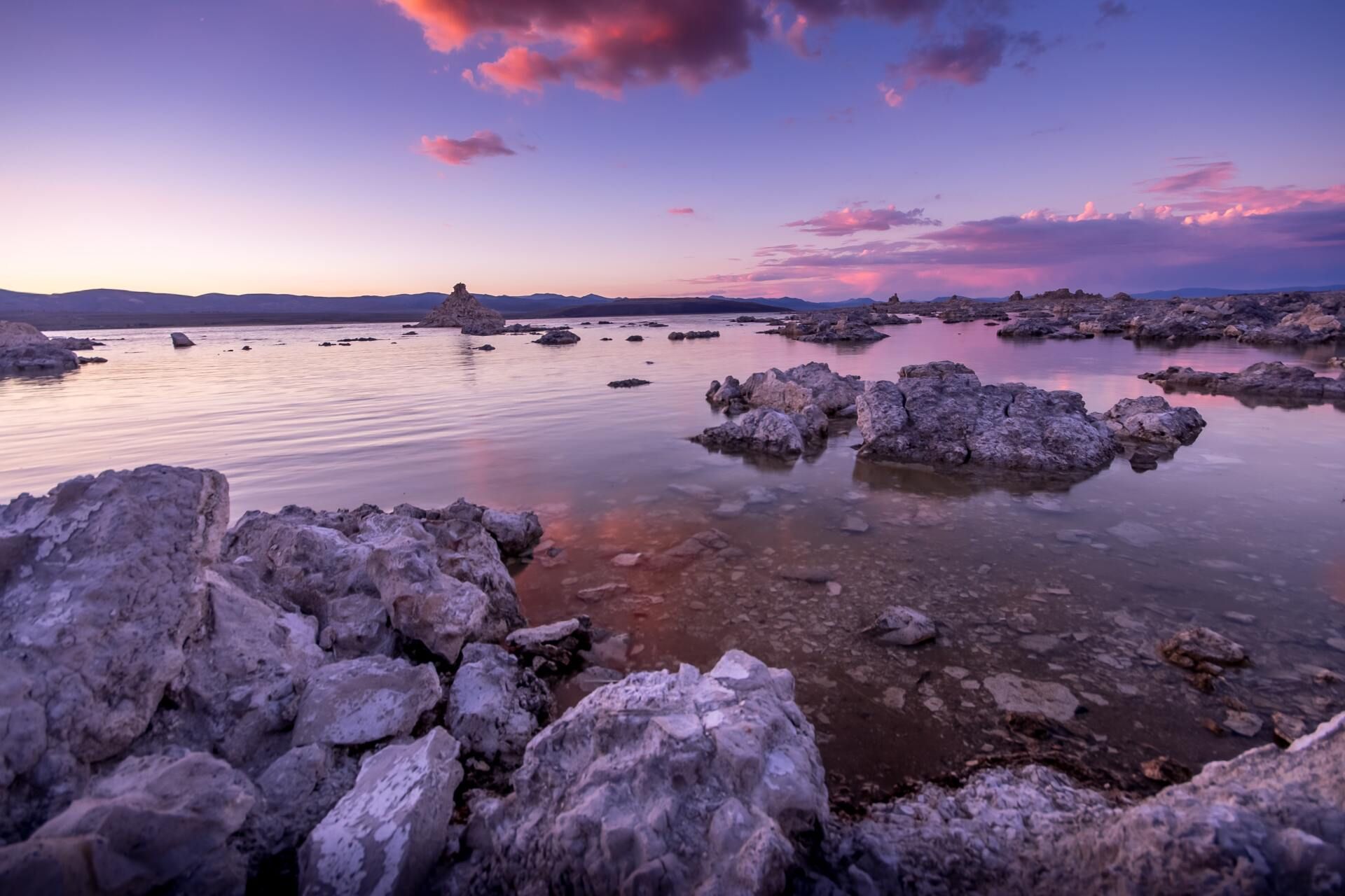 Rocky shoreline at sunset with calm water, pink-purple clouds, and distant boats on the horizon