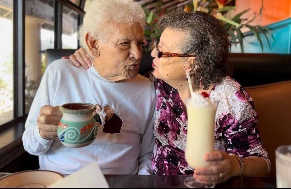 Elderly couple sharing drinks at a café, smiling and leaning close together.