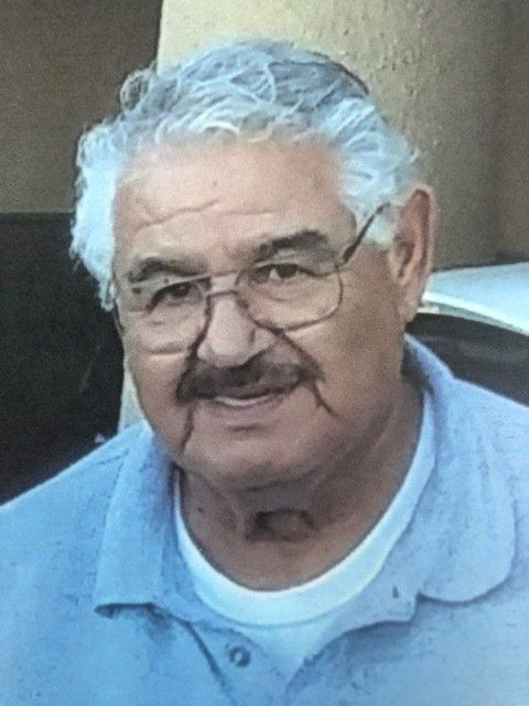 Smiling man with white hair, glasses, and a mustache wearing a light blue shirt indoors