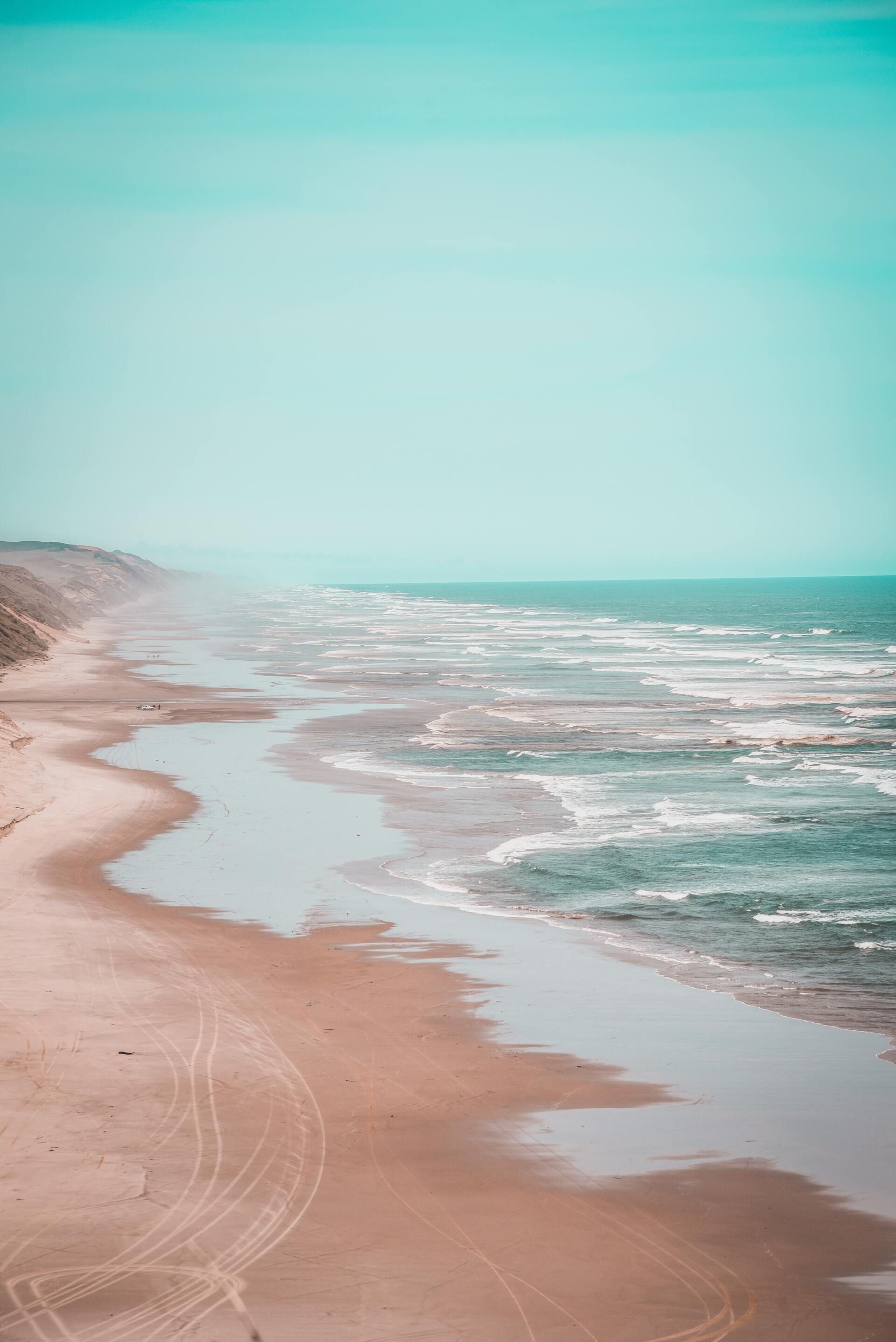 Sandy beach with gentle waves and turquoise sky, viewed along a quiet shoreline