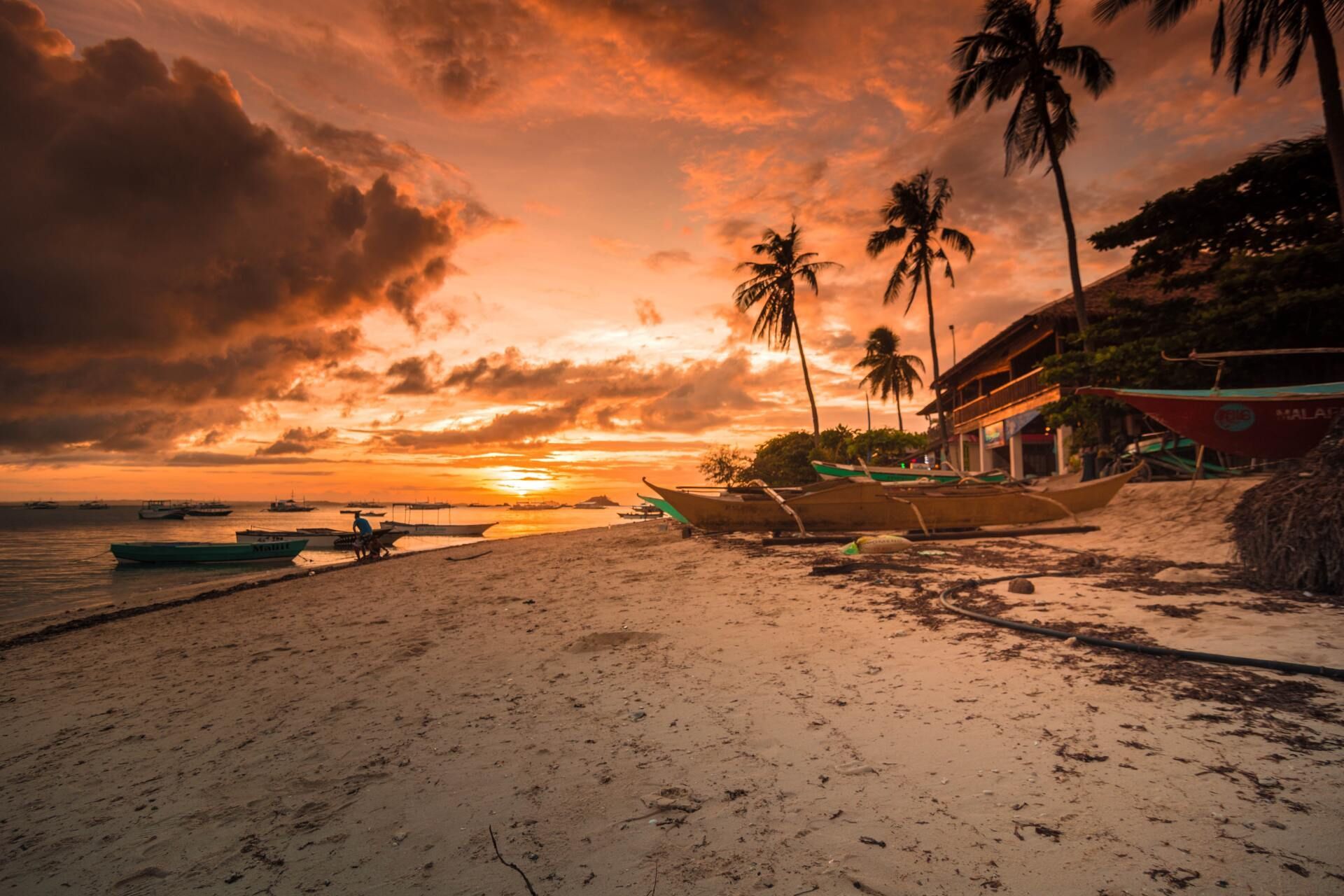 Sunset over a tropical beach with palm trees, boats, and a dramatic orange sky
