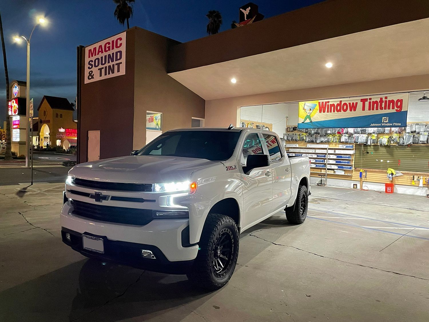 White pickup truck parked outside a tint shop at night with illuminated storefront signs.