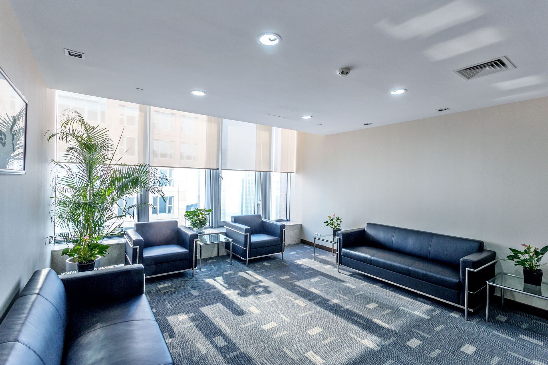 Modern waiting area with dark blue couches, armchairs, plants, and large windows with blinds.