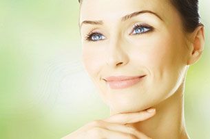 A person with a clear complexion resting their chin on their hand, smiling softly against a blurred, bright green background.