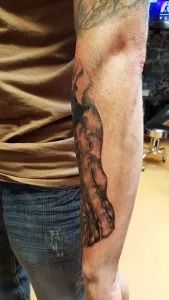 Close-up of a tattooed forearm beside blue jeans, showing a dark ink design on the outer arm.