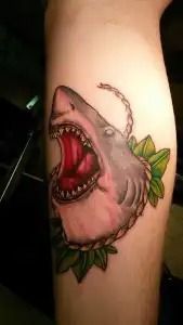 Shark head tattoo with open mouth and green leaves on a person’s forearm