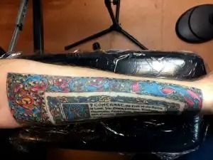 Tattooed forearm resting on a black cushion, showing a colorful rectangular tattoo design.