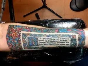 A forearm with a colorful tattoo and a rectangular text label on a black chair.