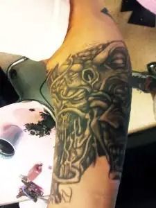 Arm tattoo of a stylized face in black and green ink, shown on an arm resting near a white object.