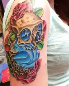 Colorful skull tattoo with blue face covering, pink roses, and green leaves on upper arm
