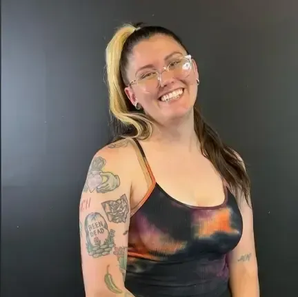 Smiling person with glasses and colorful tattoos posing against a dark gray background