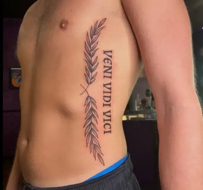 Side torso tattoo with laurel branches and the words “VENI VIDI VICI” in black ink.
