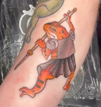 Tattoo of an orange lizard warrior holding a spear, wearing a gray outfit and skirt on a person’s leg