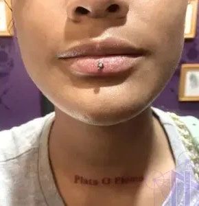 Close-up of a person’s lower face with a lip piercing and small neck tattoo against a purple wall.